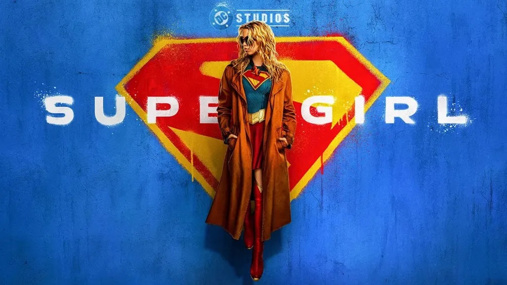 A woman in a red and blue superhero costume stands in front of a blue wall with the "DC Studios" logo, against a red and yellow superhero symbol.
