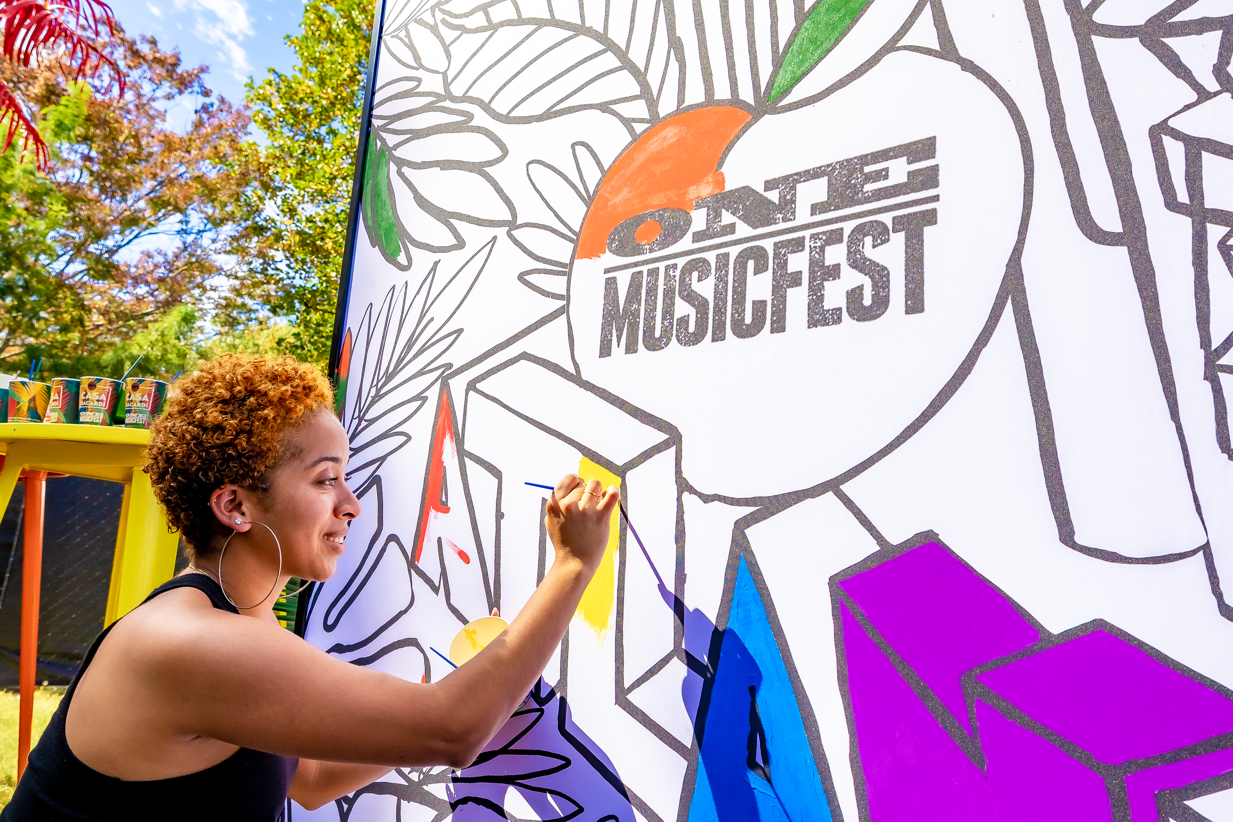 A person with curly hair painting a large mural with the text "One Musicfest" on a colorful background.