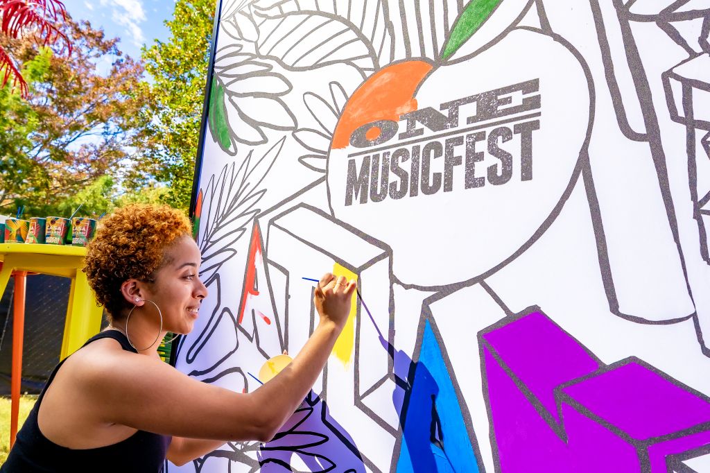 A person with curly hair painting a large mural with the text "One Musicfest" on a colorful background.