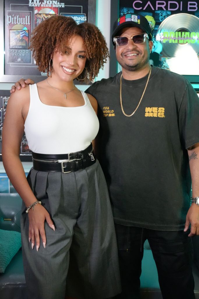 A smiling woman with curly hair and a man wearing sunglasses and a "We Are One" t-shirt stand together in front of music album covers.