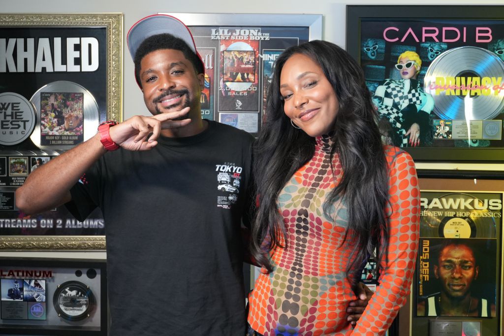 Two people, a man and a woman, posing together in front of music and entertainment memorabilia. The man is wearing a black t-shirt and the woman is wearing a colorful patterned dress. The background includes framed album covers and posters for musical artists.
