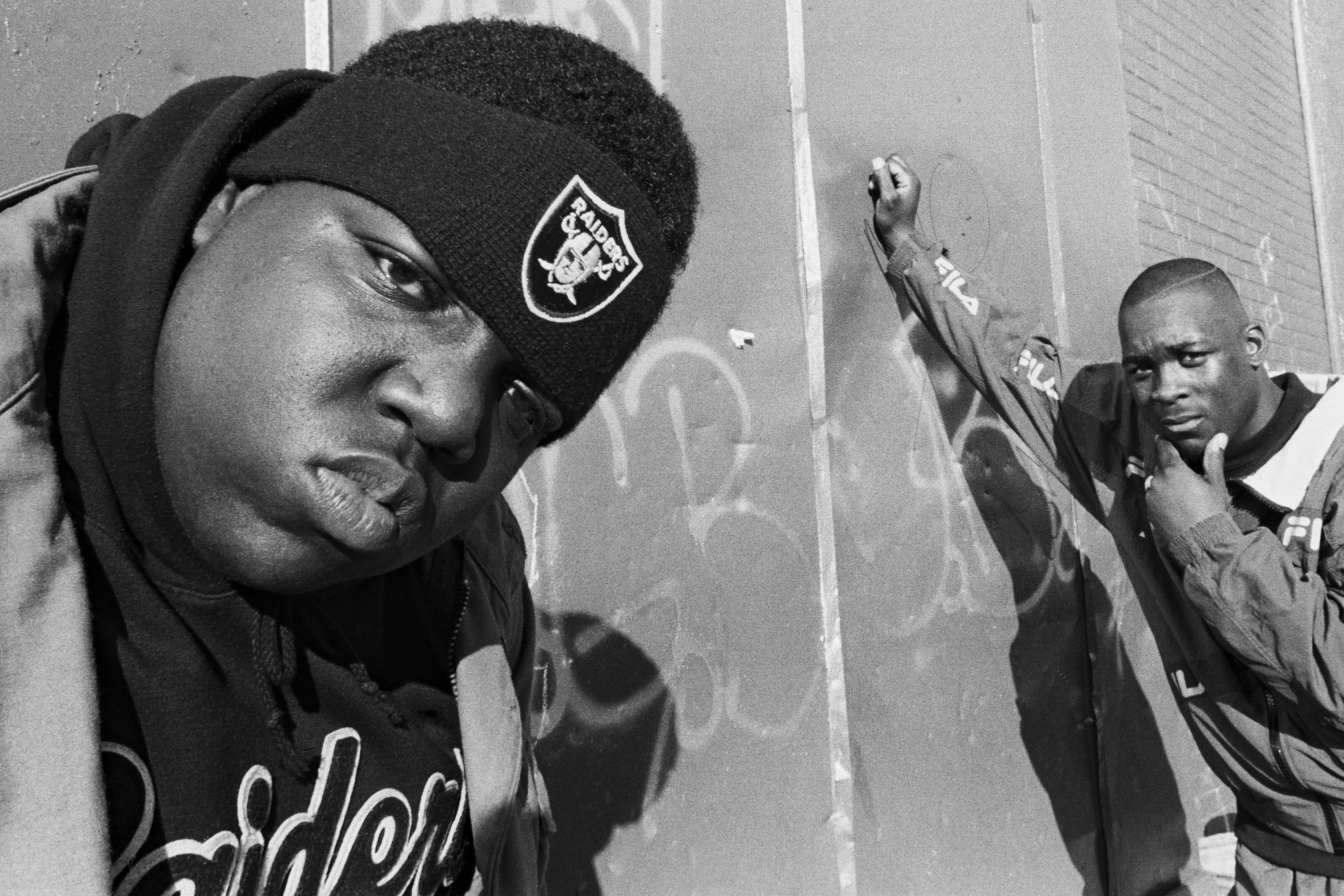 Two Black men in a dimly lit alley, one wearing a black hat with a Raiders logo.
