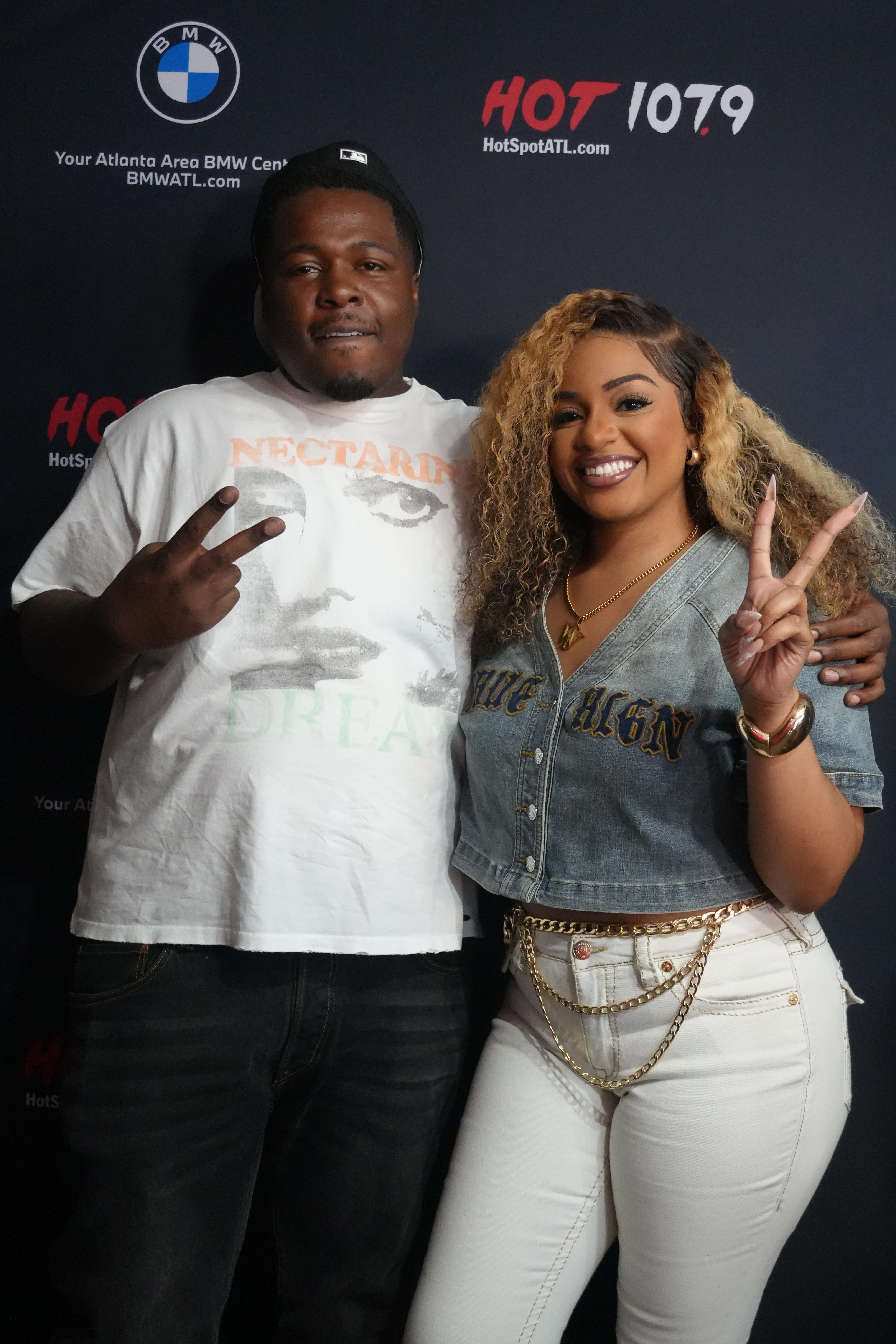 Two people posing for a photo in front of a "Hot 107.9" backdrop, with the man wearing a "Nectar" t-shirt and the woman making a peace sign.