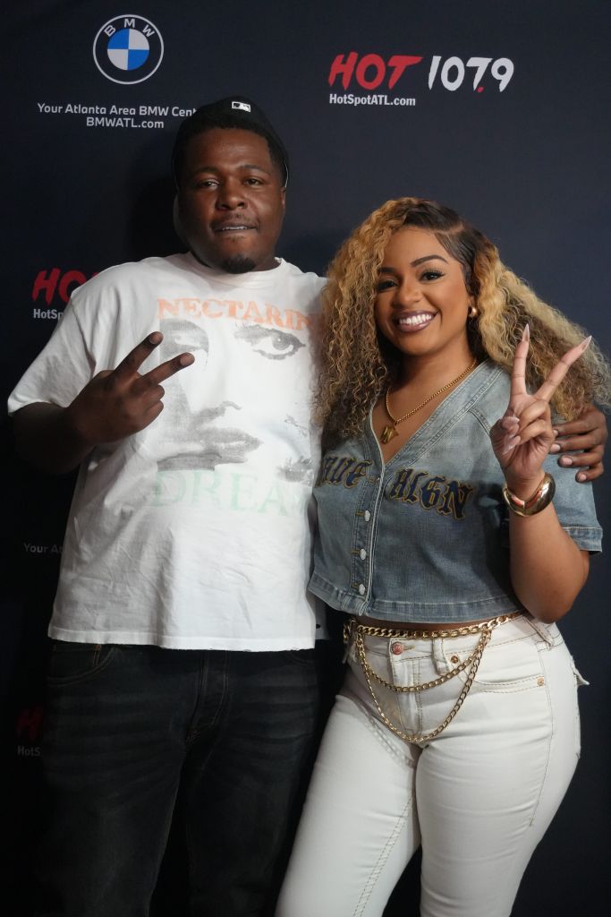 Two people posing for a photo in front of a "Hot 107.9" backdrop, with the man wearing a "Nectar" t-shirt and the woman making a peace sign.