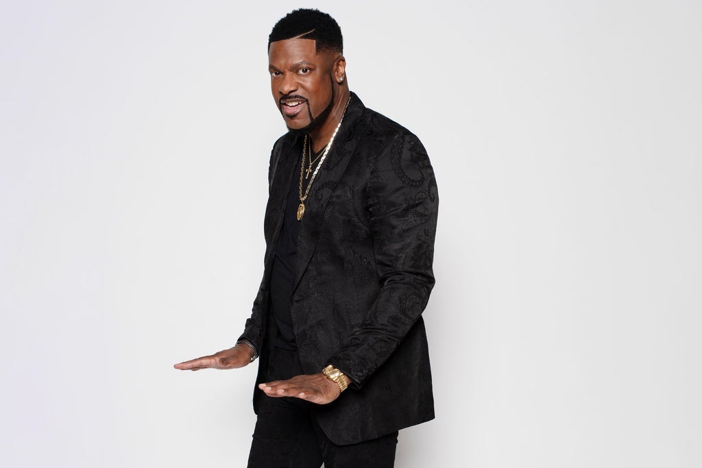 A Black man wearing a black suit and gold jewelry, standing with his arms outstretched in a confident pose.